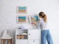 Mujer cuelga cuadros de paisajes marinos en pared de ladrillo blanco. Aprende como colocar cuadros de diferentes tamaños para un diseño armonioso.
