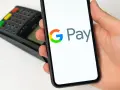 Dłoń trzyma smartfon z widocznym logo Google Pay. W tle terminal płatniczy. Szybkie i wygodne płatności.