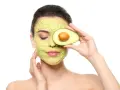 Frau mit Avocado-Gesichtsmaske und Avocado-Scheibe vor dem Auge. Die Avocado-Wirkung auf die Haut ist bekannt für ihre pflegenden Eigenschaften.
