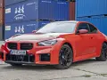 Pomarańczowe BMW M2 z czarnymi felgami, na tle niebieskich kontenerów. Sprawdź, jaka jest aktualna bmw m2 cena.