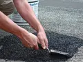 Händer som reparerar asfalt med en spade. En hink med "Asphalt" syns i bakgrunden.