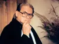 Rafał Dajbor w okularach, z dłonią na brodzie, w czarnym stroju, na tle drewnianej boazerii i bukietu suszonych traw.