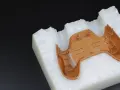 A brown plastic part, likely from a game controller, is placed on a white, textured material. This setup suggests a step in the polyurethane vacuum casting process, where a mold is being prepared.