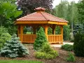Drewniana altana z czerwonym, ceramicznym dachem, otoczona bujną zielenią. Idealne miejsce na relaks, a dach z dachówki to świetny pomysł, czym pokryć dach altany tanio i estetycznie.