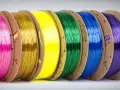 Rzędy szpul z kolorowymi filamentami: różowy, złoty, żółty, zielony, niebieski, fioletowy i szary. Różnorodne rodzaje filamentów do druku 3D.
