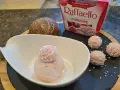 Ein Löffel Raffaello Himbeer Eis in einer Schale, daneben drei Raffaello-Kugeln und eine Kokosnuss.