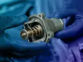 Hands in blue gloves hold a car thermostat, ready for replacement. The internal spring and valve are visible.