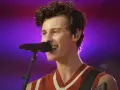 Shawn Mendes śpiewa swoje utwory na koncercie, z mikrofonem przed ustami i gitarą na ramieniu.