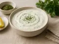 A bowl of creamy ranch dressing with herbs, a lemon wedge, and a small dish of dried herbs.