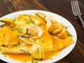 Merluza a la marinera con almejas, servida en un plato blanco sobre una mesa de madera oscura.