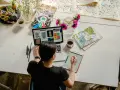 Kobieta pracuje online w domu, projektując na laptopie i tablecie. Na biurku kwiaty, kubek kawy i czaszki z pisakami.