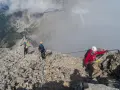 Grupa turystów pokonuje via ferrata polska. Wspinają się po skalistym grzbiecie, asekurowani linami. W tle widać góry i budynek na szczycie.