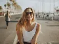 Glückliche junge Frau mit Sonnenbrille und weißem Top genießt ihr Praktikum in Spanien am Hafen.