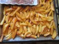Gotowe do pieczenia frytki w piekarniku, pokrojone w łódeczki i doprawione przyprawami, leżą na blasze wyłożonej papierem.