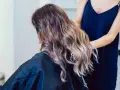 Kobieta z falowanymi włosami w odcieniach brązu i subtelnym balejażem blond. Fryzjerka poprawia jej fryzurę.