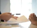 Dwie osoby przy stole, jedna podaje dokumenty, druga z dłońmi złożonymi w geście oczekiwania. Czy umowa zlecenie wlicza się do emerytury?
