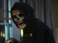 Eine Person mit Totenkopfmaske und Kapuze hält blutige Gegenstände.
