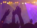 Des mains levées dans la foule d'une boîte de nuit, sous des lumières violettes et jaunes. Le son fort peut causer un acouphène après la boîte de nuit.