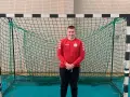 Dawid Kozak w czerwonej bluzie z długim rękawem i czarnych spodniach stoi przed bramką do piłki ręcznej.