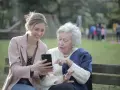 Młoda kobieta pokazuje coś na telefonie starszej pani. To scena z życia, jak z kursu opiekuna osoby starszej, gdzie uczymy się wspierać seniorów w codzienności.