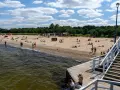 Słoneczny dzień nad morzem bałtyckim. Ludzie odpoczywają na piaszczystej plaży, niektórzy kąpią się w wodzie. Drewniane molo prowadzi na plażę.
