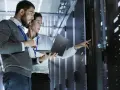 Two IT professionals collaborate on network management, one holding a laptop while the other points to a server rack.