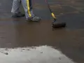 Pracownik wykonuje hydroizolację chudziaka, nakładając masę bitumiczną wałkiem.