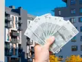 Dłoń trzyma banknoty 100 zł na tle nowoczesnych budynków mieszkalnych. Symbolizuje to podatek od sprzedaży nieruchomości.