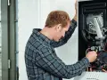 Tecnico che controlla lo scambiatore di calore della caldaia, con cavi e tubi visibili all'interno.