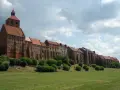 Długi, ceglany zamek w Grudziądzu w jeden dzień. Zielona trawa i krzewy przed zabytkową budowlą.
