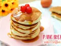 Pancakes con albumi, soffici e dorati, guarniti con sciroppo e ciliegie rosse, su un piatto rosa.