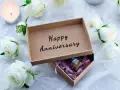 Otwarta szkatułka z napisem "Happy Anniversary" i małą buteleczką w środku, otoczona białymi różami i płonącą świecą. To idealny prezent na 4 rocznicę ślubu.