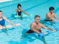 Quatre personnes font un exercice piscine avec des haltères flottants. L'eau bleue reflète la lumière du soleil.
