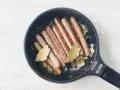 Salchichas doradas cocinándose en sartén con cebolla y hojas de laurel.