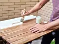 Persona pinta madera de blanco con brocha. Un frasco de pintura blanca y su tapa están cerca.