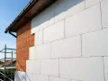 Budowa domu w toku: biały styropian na elewację przykrywa ceglany mur. Widać rusztowanie i fragment dachu.