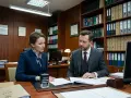 Kobieta i mężczyzna w garniturach omawiają dokumenty przy biurku. W tle regały z aktami, sugerujące sprawy dotyczące wspólności majątkowej.