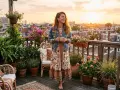 Une femme à l'allure hippie, robe fleurie et veste en jean, savoure une boisson sur un balcon fleuri, au coucher du soleil.