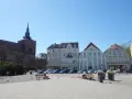Słupsk, stary rynek: kościół z wieżą, kolorowe kamienice, ławki, ludzie i gołębie na brukowanym placu.