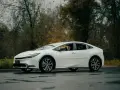Biała Toyota Prius zaparkowana na mokrej drodze, z drzewami w tle.