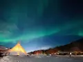 Nocne niebo nad ośnieżoną wioską, gdzie nie zachodzi słońce. Tańcząca zorza polarna nad oświetlonym tipi i domkami.