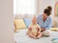 Uśmiechnięta mama wspiera dziecko, które jeszcze nie siedzi. Wspólna zabawa i ćwiczenia to klucz do rozwoju.
