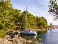 Segelboot liegt ruhig im Wasser, bereit, den Anker zu legen oder zu setzen. Grüne Bäume und Felsen säumen die Küste.