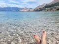 Stopy w klapkach na kamienistej plaży w **Krk National Park**. Woda krystalicznie czysta, w tle góry.