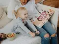 Mała dziewczynka z blond kucykami słucha bajki czytanej przez opiekunkę. Zastanawiasz się, ile zarabia opiekunka dla dziecka?