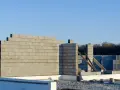 Budowa domu w toku. Wzniesiona ściana nośna z bloczków betonowych, w tle kolejne elementy konstrukcji i las.