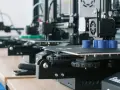 Drukarki 3D pracują, tworząc niebieskie cylindry. W tle widać kolejne maszyny, a na biurku leży smartfon, gotowy do zarządzania drukiem przez Raspberry Pi z OctoPrint.
