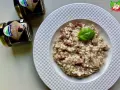 Aksamitne risotto z kawałkami kiełbasy, udekorowane listkiem bazylii. Obok stoją słoiczki z sosem truflowym, sugerujące jego obecność w daniu.