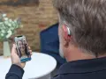 Mężczyzna z aparatem słuchowym rozmawia przez wideorozmowę na telefonie. Aplikacja na telefon pozwala na zdalne dopasowanie aparatów.