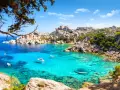 Turkusowa woda i skaliste wybrzeże Costa Smeralda na Sardynii. Kilka łodzi kołysze się na falach.